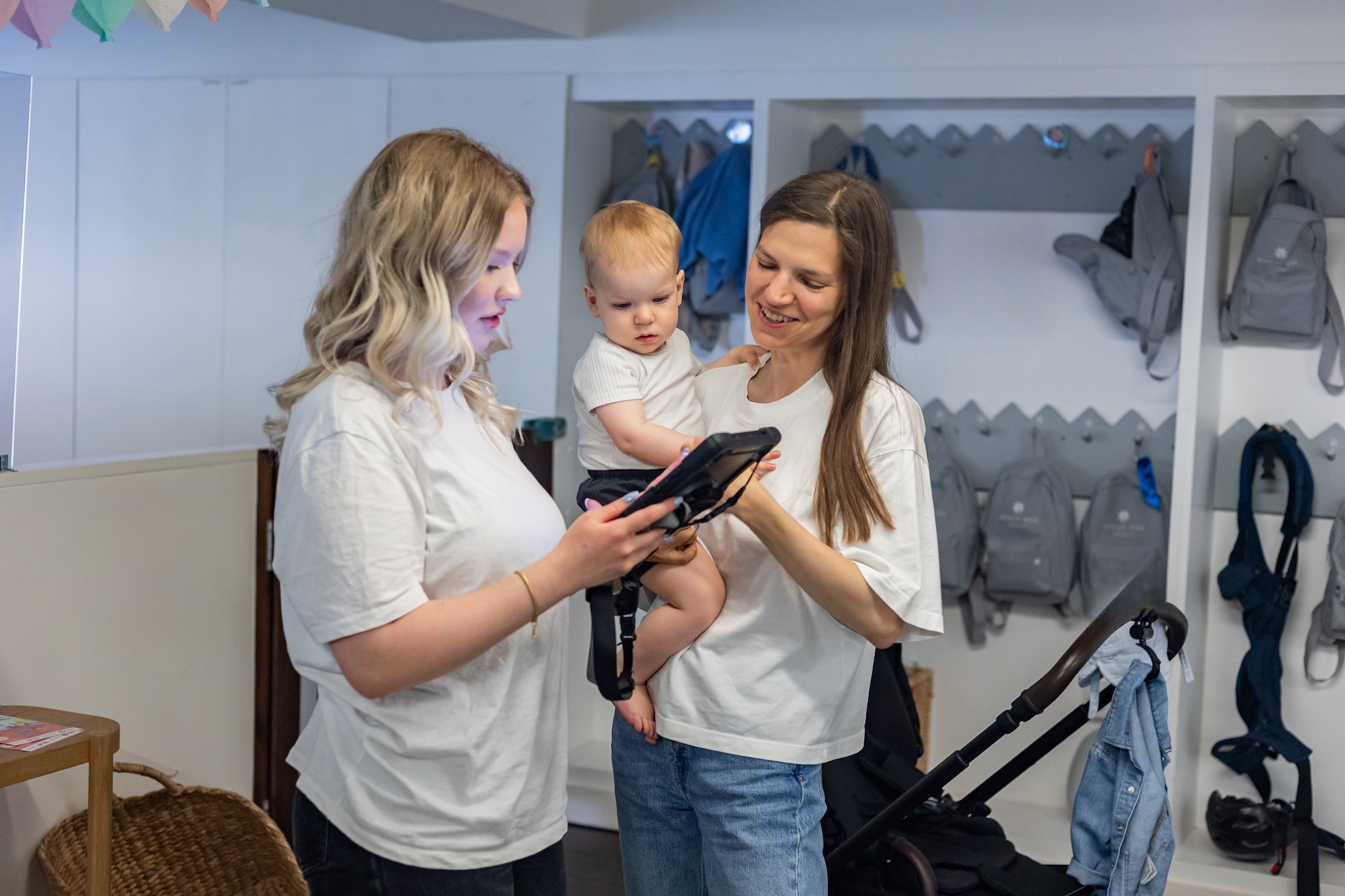 A parent and nursery educator in a warm, brief handover conversation at drop-off — relaxed, informative, and communicative. The child visibly comfortable and settled between them.