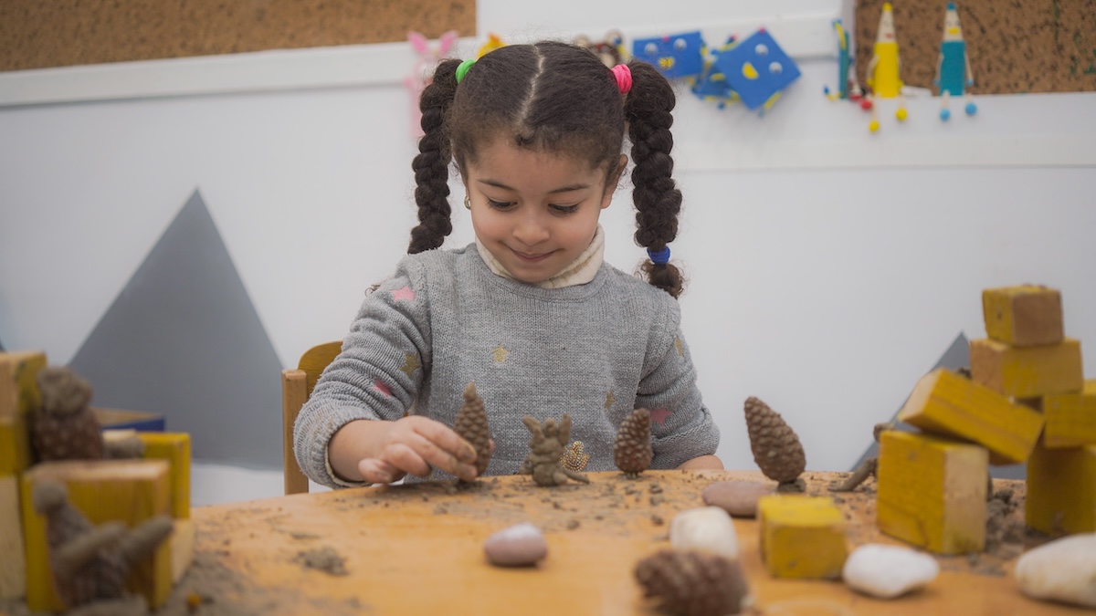 Children gathered around a nature table examining leaves, stones, and pinecones with magnifying glasses — curious expressions, hands-on exploration in an outdoor or nature-inspired indoor setting.